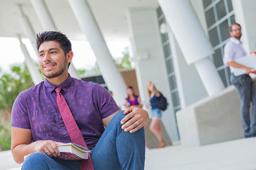 male student with book on steps of special event center